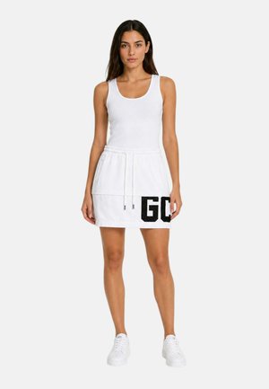 Vrouw staand, gekleed in een witte mouwloze jurk met een trekkoord in de taille en zwarte letters "GO" op de rok, gecombineerd met witte sneakers.