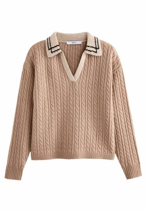 Personne portant un pull beige en maille torsadée avec un col blanc, un jean noir fuselé, des baskets noires, tenant un manteau noir sur une épaule.