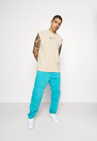 Beige ermeløs t-skjorte med logo foran, satt sammen med turkise cargo-bukser med sidelommer. Modellen har på seg hvite joggesko.