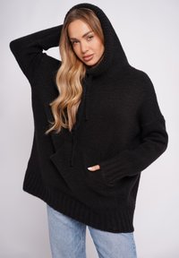 Czarna dzianinowa, luźna bluza z wysokim kołnierzem, sznurkiem ściągającym, przednią kieszenią i ściągaczem na dole, noszona na jasnoniebieskich dżinsach. Tkanina o fakturze.