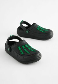 Sabots noirs avec un motif de os vert sur le dessus, présentant un intérieur en peluche et une sangle de talon réglable pour un ajustement sécurisé.