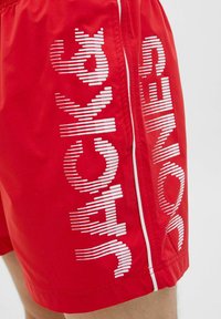 Bañador rojo con texto horizontal blanco en el lateral, con una textura suave y detalles en la costura lateral. El texto dice "NADAR" y "CHALECOS."