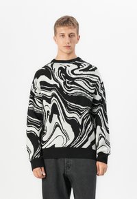 Jonge man in een zwart-wit abstract patroon sweater met zwirls en donkergrijze broek, staande tegen een eenvoudige witte achtergrond.