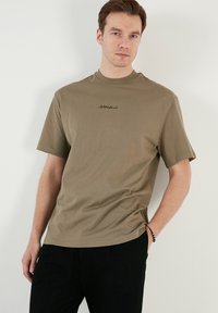 Oliven grøn bomulds t-shirt med korte ærmer, rund hals og sort broderet tekst på brystet. Løs, afslappet pasform og glat tekstur.