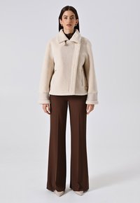 Giacca in bouclé crema con colletto, tasche laterali e polsini a contrasto, abbinata a pantaloni marroni a vita alta. Texture liscia.