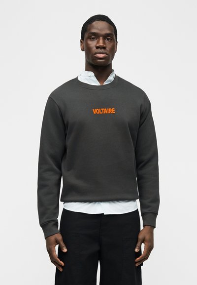 Μαύρος άνδρας που φοράει γκρι σκοτεινό φούτερ με τη λέξη «VOLTAIRE» σε πορτοκαλί στο στήθος, λευκό πουκάμισο από μέσα και μαύρο παντελόνι, στέκεται ακίνητος.