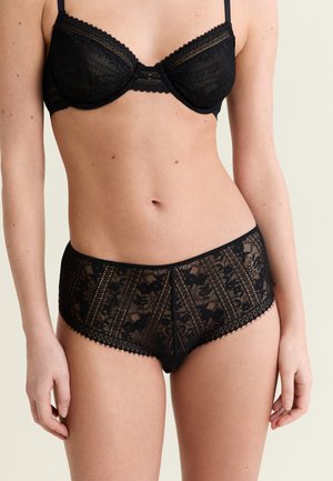 Woman wearing black lace lingerie set with underwire bra and high-waist patterned lace panties, posed against plain light background.