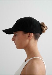Gorra de béisbol de ante negro con visera curva, que presenta una superficie texturizada y un acento de botón en la parte superior. El cabello está peinado en un moño en la parte posterior.