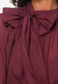 Detaliu blouse burgundy, cu un mare fundă la gât, realizată dintr-un material ușor și fluid, cu o textură moale și drapată.