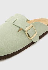 Zelena nubuk slip-on cipela s okruglim vrhom, s zlatnim akcentom kopče i smešnim gumenim đonom. Glatka tekstura i minimalizam u dizajnu.