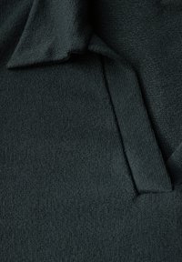 Mörkgrön stickad plagg med en slät textur, med en vikbar krage och synliga stygn i halsringningen och kanterna.