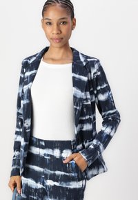 Marineblauwe en witte tie-dye patroon blazer met een zachte textuur, gecombineerd met een witte getailleerde top. Model in een casual houding met handen in zijn zakken.