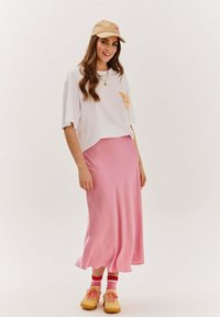 Zkrátené biele tričko s grafickým dizajnom, kombinované s ružovou saténovou midi sukňou. Béžová čiapka a žlté tenisky s červenými a ružovými pruhovanými ponožkami.