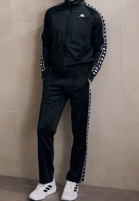 Personne portant un survêtement noir Kappa avec des bandes blanches à logo sur les manches et les jambes du pantalon, assorti à des baskets blanches Adidas.