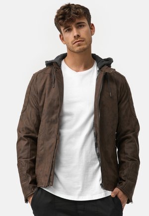 Braune Lederjacke mit grauem Kapuzen, figurnaher Schnitt, Reißverschluss vorne, strukturierte Oberfläche und Schulterakzente. Getragen über einem weißen T-Shirt.