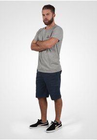 Sivá bavlnená tričko s malým čiernym logom, skombinované s námorníckymi cargo šortkami. Čierne tenisky so zlatým akcentom dotvárajú outfit.