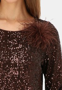 Brązowy top z cekinami, z okrągłym dekoltem, długimi rękawami oraz teksturowanym akcentem z piór na lewym ramieniu.
