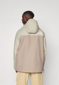 Beige og lys tan hettejakke med en glatt tekstur og logo på baksiden, med en avslappet passform og justerbar hette.