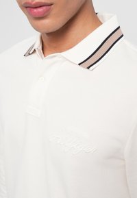 Camisa polo branca feita de tecido texturizado, com uma pala de três botões e um colarinho riscado com acentos em bege e azul. Logotipo bordado no peito.