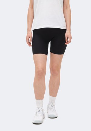 Osoba ubrana w czarne dopasowane spodenki rowerowe, białe skarpetki sportowe, białe trampki oraz luźną białą koszulkę, stojąca na jednolitym tle.