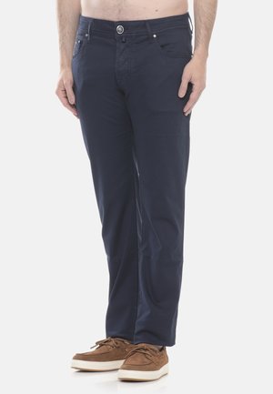 Uomo con pantaloni blu navy e scarpe casual marroni, in piedi con le mani rilassate lungo i fianchi, su sfondo bianco.