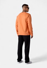 Langärmliges oranges Shirt aus Baumwolle, kombiniert mit schwarzen Hosen und weißen Sneakers. Das Shirt hat ein einfaches Design mit einem Rundhalsausschnitt.