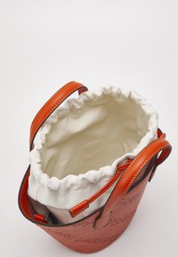 Sac seau en cuir marron avec motif perforé, doté d'une fermeture à cordon en tissu blanc et de poignées en cuir assorties.