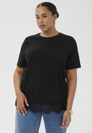 Femme aux cheveux foncés attachés en chignon portant un haut noir à manches courtes avec ourlet en dentelle et un jean bleu, posant devant un fond clair uni.