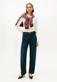 Maglia bianca a maniche lunghe, jeans a gamba larga blu scuro, cintura nera e un maglione a righe bordeaux appoggiato sulle spalle. Tacchi neri a punta.