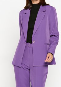 Lila skräddarsydd kavaj med slagkrage, enkel knappstängning och två sidofickor, matchad med byxor i samma färg och en svart ribbad topp.
