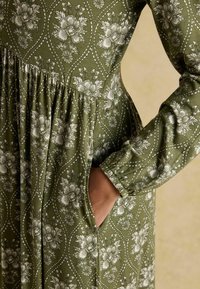 Zelené šaty s kvetinovým vzorom, dlhými rukávmi, zúženým pásom a bočnými vreckami. Materiál má hladkú textúru a prepracované biele detaily.