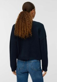 Sieviete ar gariem, viļņainiem matiem, kas savīti astē, valkā tumšu džemperi un zilos džinsus, redzama no aizmugures uz neitrāla fona.