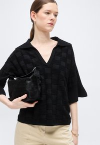 Femme à la peau claire et aux cheveux bruns attachés en queue de cheval basse, portant un haut noir texturé à col en V et un pantalon beige, tenant un petit sac à main noir.