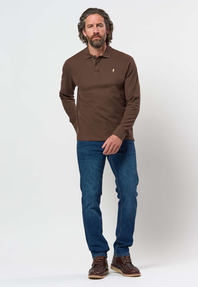 Man met krullend haar en baard, dragend een bruin poloshirt met lange mouwen, blauwe spijkerbroek en bruine leren laarzen, staand tegen een effen achtergrond.