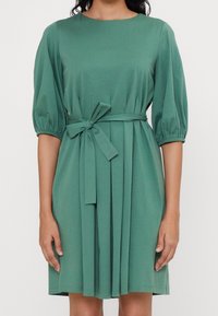Frau in einem knielangen grünen Kleid mit dreiviertel Puffärmeln und einem Taillenknoten, die vor einem schlichten Hintergrund steht.
