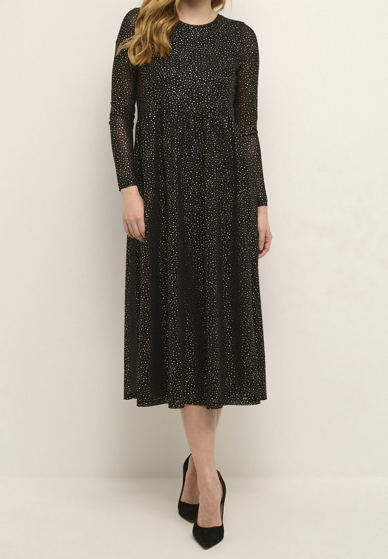 Femme portant une longue robe noire à petits pois blancs, manches longues, et des talons hauts noirs pointus, debout contre un mur uni.