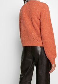 Sweater orange doux et texturé avec un col rond et des manches légèrement bouffantes. Associe avec un pantalon en cuir noir brillant, mettant en valeur un design moderne.
