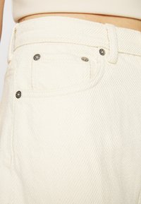 Tecido branco texturizado com uma tecelagem diagonal visível. Apresenta um pequeno bolso com rebites de metal e uma cintura resistente.