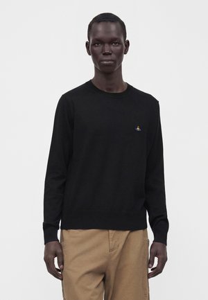 Man met kort haar die een zwart shirt met lange mouwen draagt met een klein kleurrijk embleem op de borst en beige broek, staand tegen een witte achtergrond.