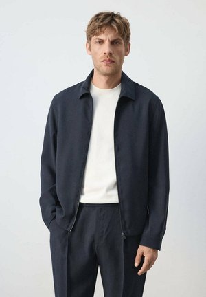 Hombre con chaqueta azul marino sobre camisa blanca y pantalones azul marino, de pie con una mano en el bolsillo contra un fondo liso.