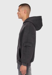 Férfi sötétszürke, oversized kapucnis pulóverben, amelynek nagy kapucnija és zsebfedél részlete van az ujjon, profillal áll egy egyszínű háttér előtt.