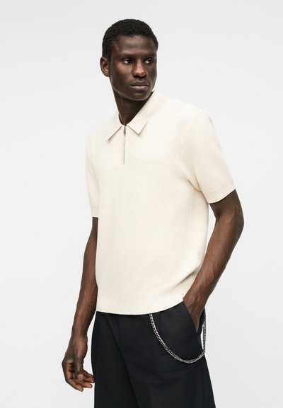 Jeune homme portant un polo en maille crème à manches courtes avec une fermeture éclair et un pantalon noir avec une chaîne argentée, debout devant un fond clair uni.