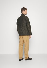 Dark green, lightweight jacket with a high collar, textured finish, and zippered side pockets; paired with beige trousers and black shoes.