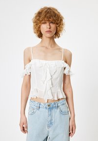 Weißes Off-Shoulder-Oberteil mit rüschenbesetzten Kanten, einer Schnüraausschnitt an der Vorderseite und dünnen Trägern, kombiniert mit hellblauen, ausgewaschenen Jeans.