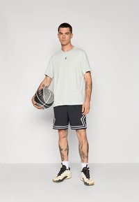 Lysegrå t-skjorte med svart logo, svarte atletiske shorts med hvite detaljer, og svarte og kremfargede basketballsko. Holder en basketball med mønster.