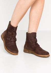 Jalkoja, joissa on tummanruskeat nahkaiset nilkkasaappaat, joissa on nauhat ja vaaleanruskeat ommellut pohjat, valkoisella lattialla.