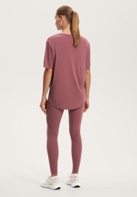 Halenka s krátkym rukávom v mauve farbe s ladiacimi legínami, ktoré majú jemné švy. Model nosí biele a krémové bežecké topánky.