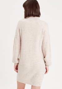 Robe-pull tricotée en beige clair, avec manches longues, col montant et coupe décontractée, présentant une texture visible et des poignets côtelés.