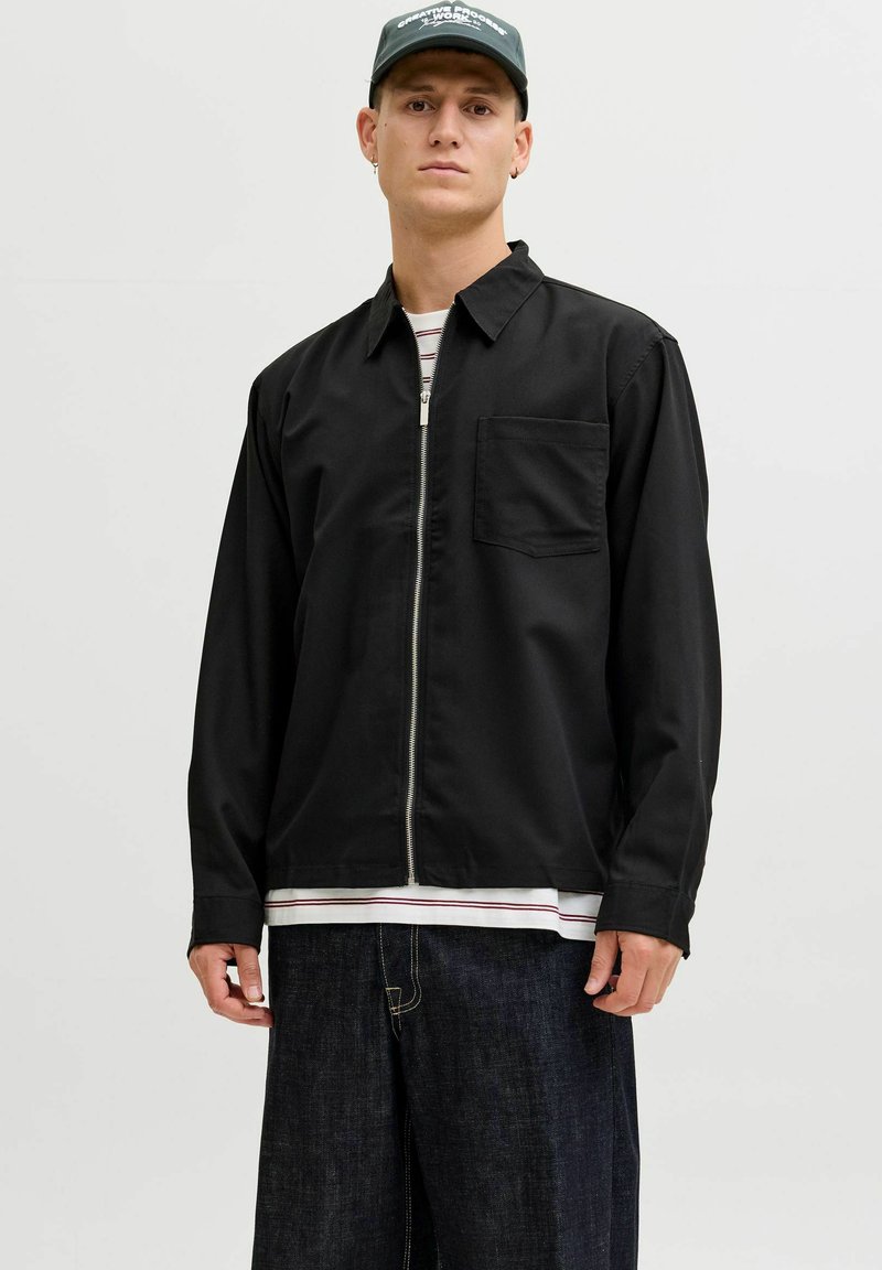 Giovane uomo che indossa una giacca nera con cerniera, una maglietta a righe sotto, jeans larghi scuri e un cappello verde scuro con scritta bianca.