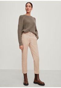 Pantaloni cargo beige chiaro con tasche laterali abbinati a un maglione in maglia marrone testurizzata. Stivaletti alla caviglia marroni intagliati. Sfondo neutro.
